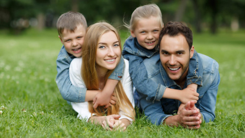 Two smiling parents lying on grass with two young children on their backs outdoors